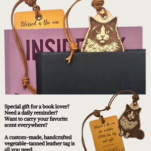 以下が含まれることがあります： 猫のイラストと「Blessed is the one」の文字が入った革製のブックマーク。ブックマークは茶色の革紐で取り付けられています。1つのブックマークは本に挿入されています。もう1つのブックマークは本の隣にあります。