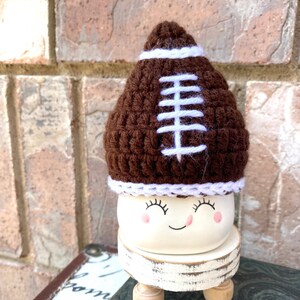 May include: A white ceramic planter with a happy face and closed eyes. The planter is wearing a brown crocheted football hat with white stitching.