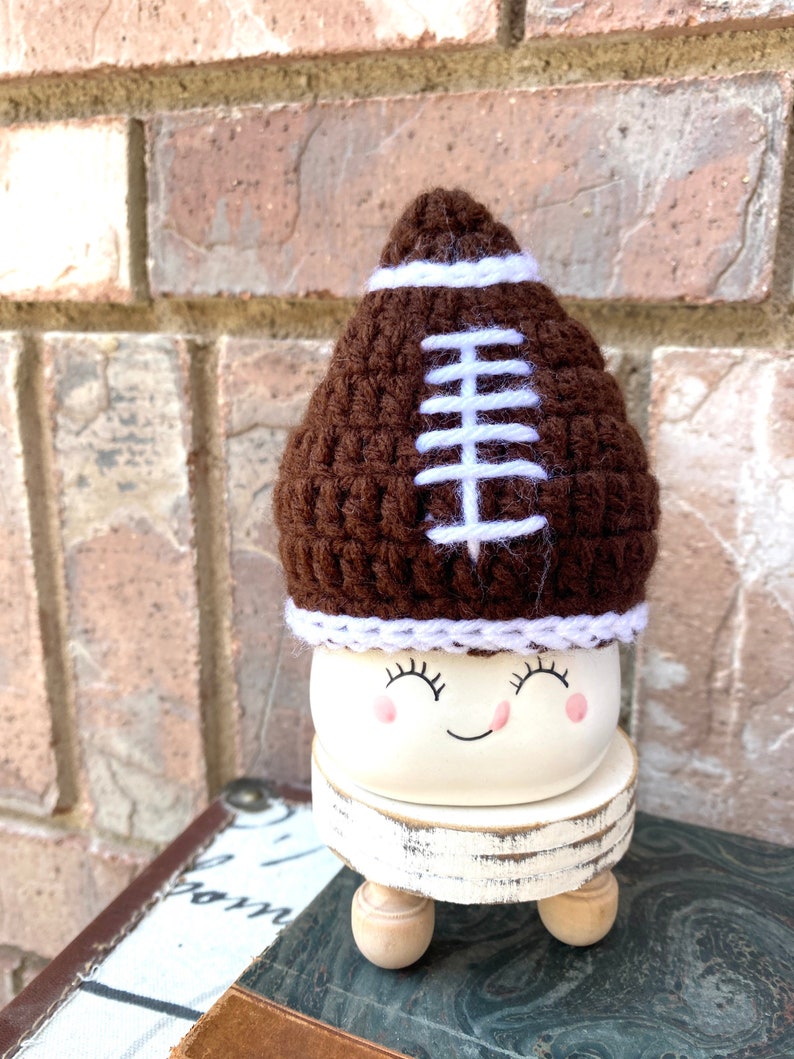 May include: A white ceramic planter with a happy face and closed eyes. The planter is wearing a brown crocheted football hat with white stitching.
