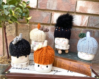 Könnte beinhalten: Eine Sammlung von Marshmallow-Figuren aus Keramik, jede mit einem einzigartigen, handgehäkelten Hut. Die Hüte sind in verschiedenen Farben erhältlich, darunter Schwarz, Orange, Creme und Grau, einige mit dekorativen Akzenten. Die Figuren werden auf kleinen Holzständern präsentiert.