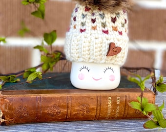 May include: A white ceramic mug with a smiling face and closed eyes, wearing a white knitted hat with a brown pom-pom and a small wooden heart. The mug is sitting on top of an old, brown leather-bound book.