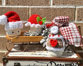 Puede incluir: Tres tazas de cerámica blanca con caras sonrientes están decoradas con gorros de Navidad de ganchillo. Las tazas están sentadas en un trineo de madera, que está encima de una maleta vintage. Los gorros son rojos, verdes y marrones con ribetes blancos.