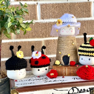 May include: A collection of handmade ceramic figurines, each adorned with a unique crocheted hat. The hats are designed to resemble a ladybug, a bee, and a beehive. The figurines are arranged on a decorative trunk, with a small potted plant in the background.