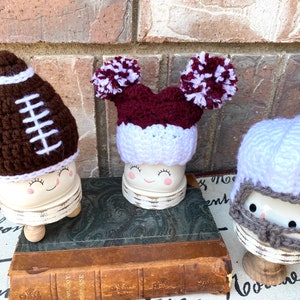 May include: Three white ceramic figurines with different crocheted hats. The first figurine has a brown and white football hat. The second figurine has a maroon and white pom-pom hat. The third figurine has a white crocheted helmet with a gray face mask.
