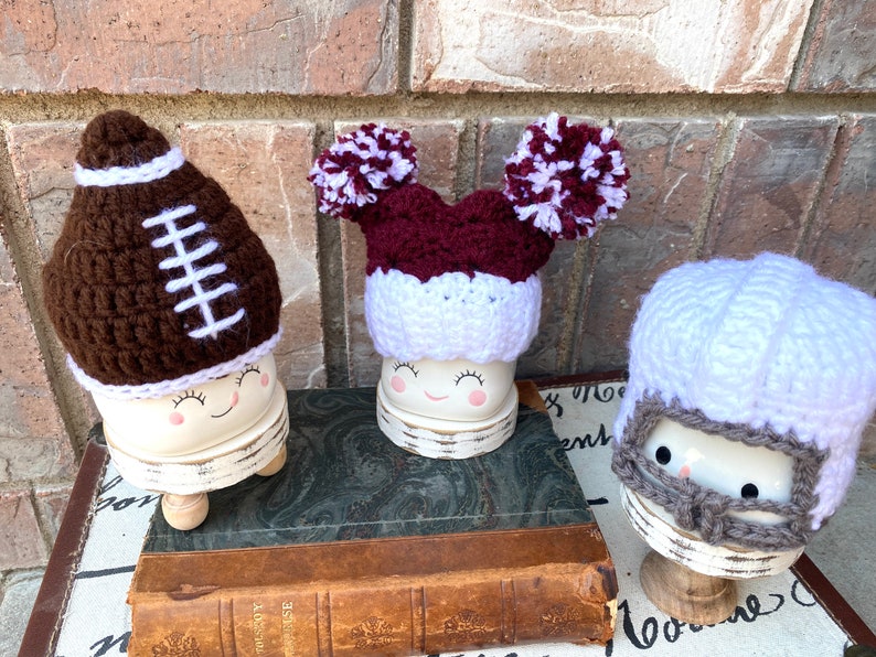 May include: Three white ceramic figurines with different crocheted hats. The first figurine has a brown and white football hat. The second figurine has a maroon and white pom-pom hat. The third figurine has a white crocheted helmet with a gray face mask.