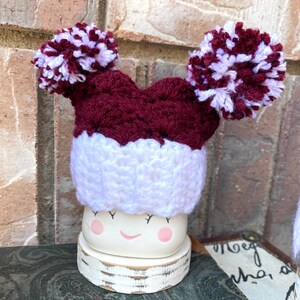 May include: A white ceramic mug with a smiling face and pink cheeks, wearing a maroon and white crocheted hat with two pom-poms.
