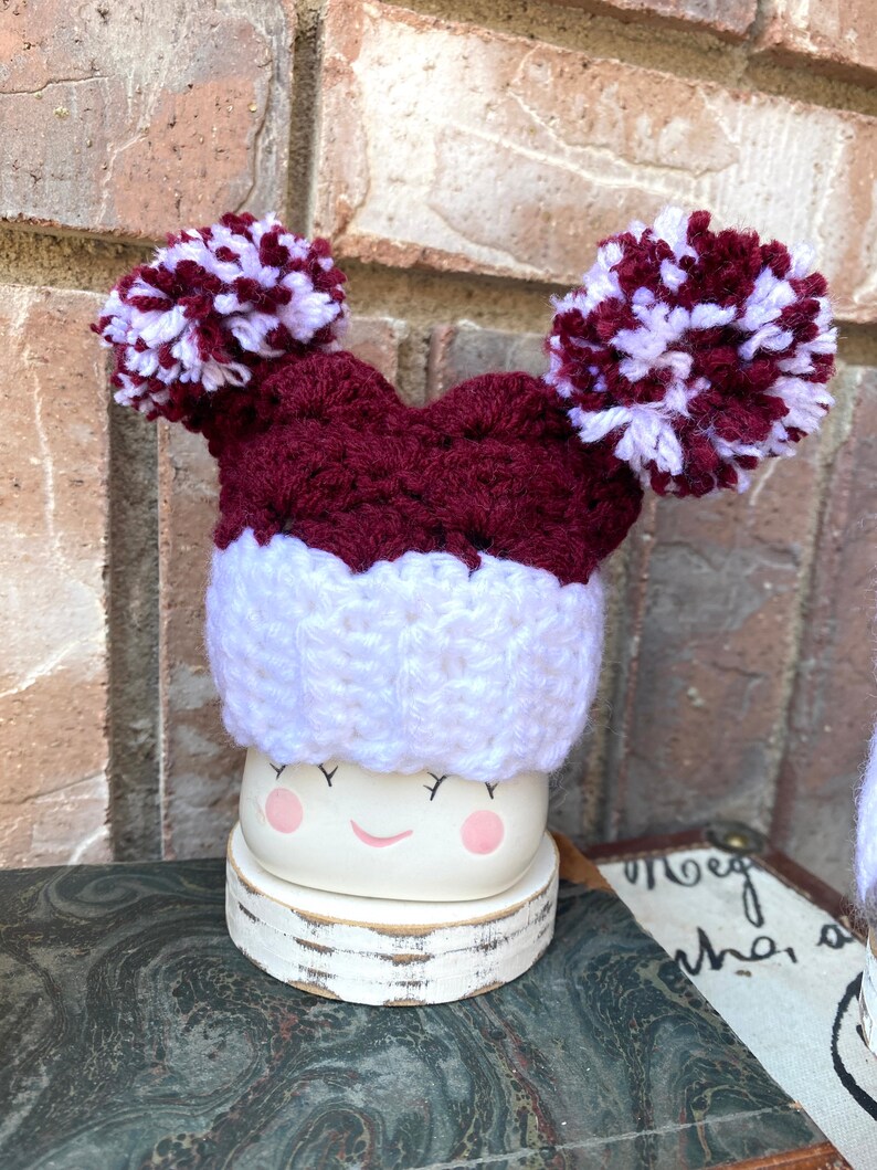 May include: A white ceramic mug with a smiling face and pink cheeks, wearing a maroon and white crocheted hat with two pom-poms.