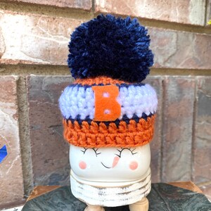 May include: A white ceramic snowman figurine with a crocheted hat in orange, blue, and white with a blue pom-pom on top. The hat has a white letter "B" on the front. The snowman has a painted smile and closed eyes.