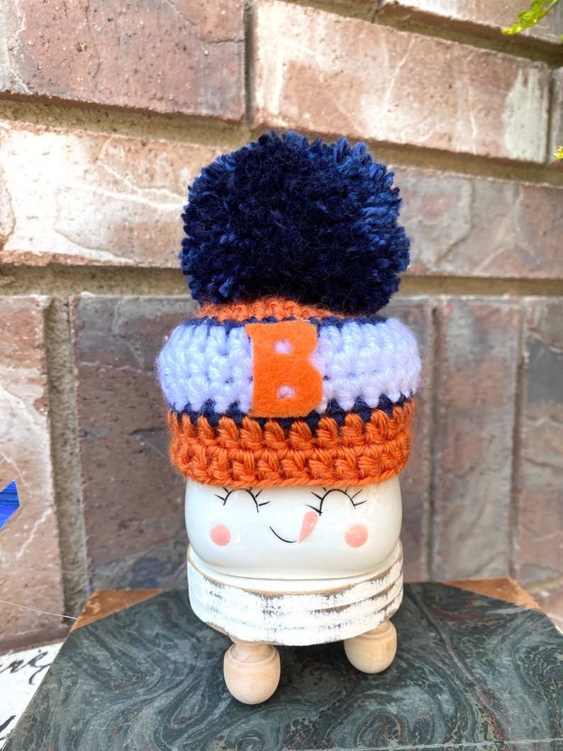 May include: A white ceramic snowman figurine with a crocheted hat in orange, blue, and white with a blue pom-pom on top. The hat has a white letter "B" on the front. The snowman has a painted smile and closed eyes.