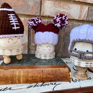 May include: Three ceramic figures with painted faces wearing crocheted hats. The hats are a brown football, a maroon and white pom-pom, and a white helmet with a gray face mask.
