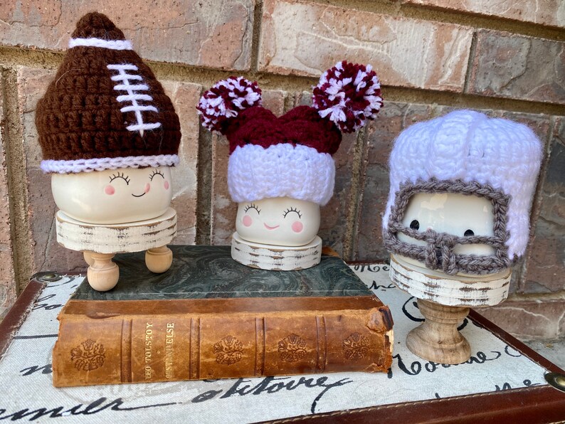 May include: Three ceramic figures with painted faces wearing crocheted hats. The hats are a brown football, a maroon and white pom-pom, and a white helmet with a gray face mask.