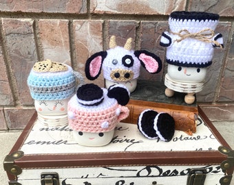 May include: Four white ceramic mugs with crocheted hats. The hats are decorated with different designs, including a cow, a cookie, and a chocolate chip cookie. The mugs are sitting on a vintage suitcase with a brown leather trim.