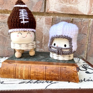 May include: Two white ceramic figurines with crocheted football helmet hats. The figurine on the left is wearing a brown and white football helmet hat and has a smiling face. The figurine on the right is wearing a white and gray football helmet hat. Both figurines are sitting on small wooden stands. The figurines are sitting on top of an old book.