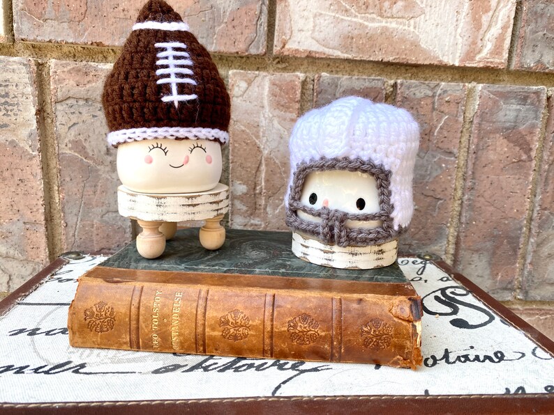 May include: Two white ceramic figurines with crocheted football helmet hats. The figurine on the left is wearing a brown and white football helmet hat and has a smiling face. The figurine on the right is wearing a white and gray football helmet hat. Both figurines are sitting on small wooden stands. The figurines are sitting on top of an old book.