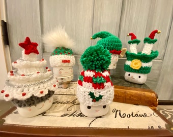 Könnte beinhalten: Eine Sammlung skurriler, handgefertigter Weihnachtsdekorationen. Die Dekorationen umfassen Figuren in Marshmallow-Form mit gehäkelten Hüten, darunter ein Weihnachtsbaum, Elfenhüte und ein Hut mit einem flauschigen weißen Bommel. Die Hüte sind rot, grün und weiß.