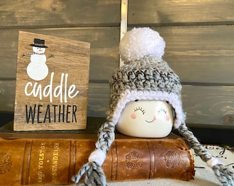 Könnte beinhalten: Eine graue und weiße Häkelmütze mit einem weißen Bommel oben. Die Mütze liegt auf einem Buch mit dem Text "cuddle weather" auf einem Holzschild dahinter.