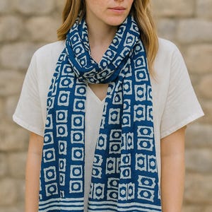 Peut inclure: Écharpe à motifs bleus et blancs. Le motif répété est composé de carrés sur fond bleu foncé et de formes blanches. La personne porte un haut blanc à col en V. L'écharpe est en matière douce.