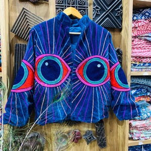 Puede incluir: Chaqueta de terciopelo azul con diseño de ojos bordados. La chaqueta presenta grandes ojos coloridos en la parte delantera y las mangas, con detalles en rojo, rosa y verde. Líneas verticales irradian de los ojos. La chaqueta se exhibe en una percha de madera.