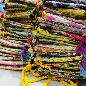 Puede incluir: Montones de tela colorida estampada con adornos y borlas decorativas. Las telas presentan una variedad de colores, incluyendo amarillo, rosa, verde y azul. Algunas telas tienen adornos dorados y detalles de cuentas amarillas.