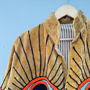 May include: A mustard-colored velvet jacket with black embroidered sunburst designs. The jacket features a mandarin collar and a striped lining. The sunburst design includes orange, turquoise, and black details, creating a striking visual contrast.