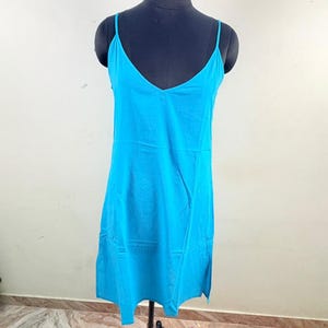 Può includere: Un abito sottoveste turchese con sottili spalline e scollo a V. L'abito è esposto su un manichino nero con un supporto in metallo. L'abito ha un design semplice e fluido.