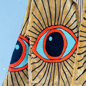 May include: A tan-colored, long-sleeved garment with large, embroidered eye designs. The eyes feature black pupils, light blue irises, and orange outlines, set against a radiating pattern of black lines. The garment appears to be made of a soft, textured fabric.