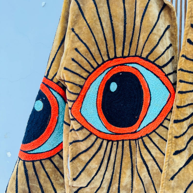 May include: A tan-colored, long-sleeved garment with large, embroidered eye designs. The eyes feature black pupils, light blue irises, and orange outlines, set against a radiating pattern of black lines. The garment appears to be made of a soft, textured fabric.