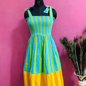 Puede incluir: Un vestido sin mangas a rayas en tonos azules, verdes y amarillos. El corpi&ntilde;o es fruncido y las correas est&aacute;n atadas a los hombros. La falda se ensancha desde la cintura. El vestido se exhibe en un maniqu&iacute; negro.