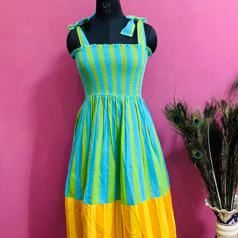 Puede incluir: Un vestido sin mangas a rayas en tonos azules, verdes y amarillos. El corpi&ntilde;o es fruncido y las correas est&aacute;n atadas a los hombros. La falda se ensancha desde la cintura. El vestido se exhibe en un maniqu&iacute; negro.