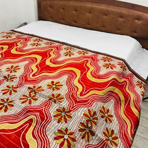 May include: A bed with a brown leather headboard and a white sheet. The bed is covered with a red, yellow, and cream-colored patterned blanket. The blanket features a floral design with wavy lines.