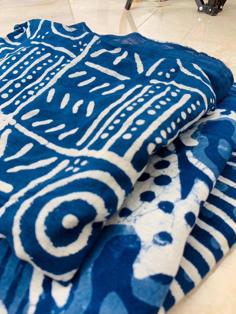 May include: Folded blue fabric with white geometric patterns. The fabric has a textured appearance and is layered, showcasing the intricate designs. The patterns include lines, dots, and circular shapes, creating a visually appealing textile.