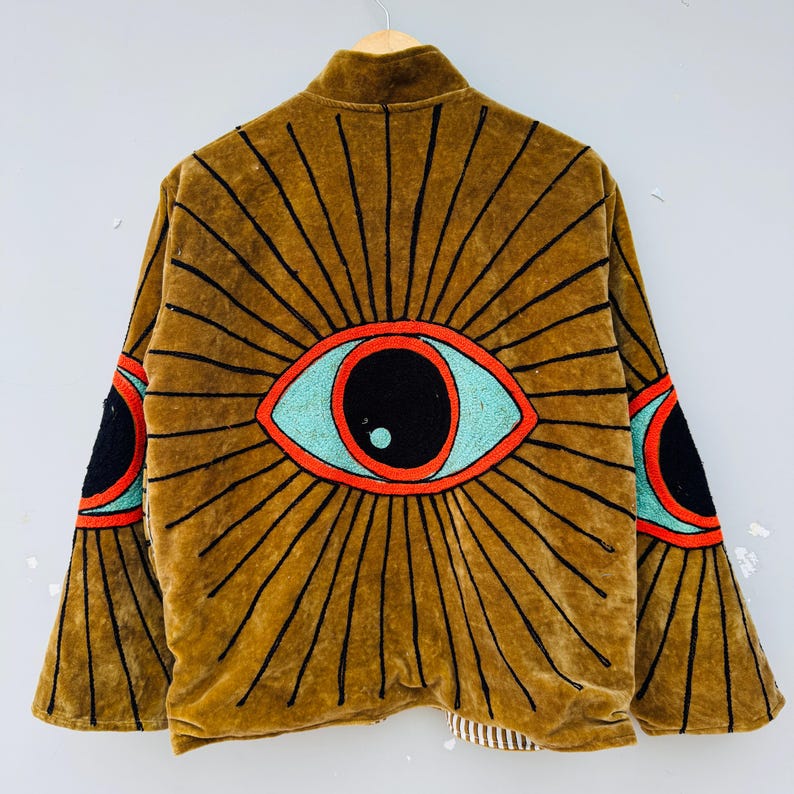 May include: A brown velvet jacket with a large eye design on the back and sleeves. The eye features a black pupil, a turquoise iris, and a red outline, with black rays emanating from it. The jacket is hanging on a wooden hanger.