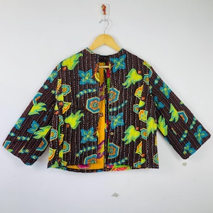 Puede incluir: Chaqueta marrón con frente abierto y un vibrante estampado floral en azul, amarillo y verde. La chaqueta es reversible, con un interior amarillo y naranja. La chaqueta está colgada de una percha de madera.