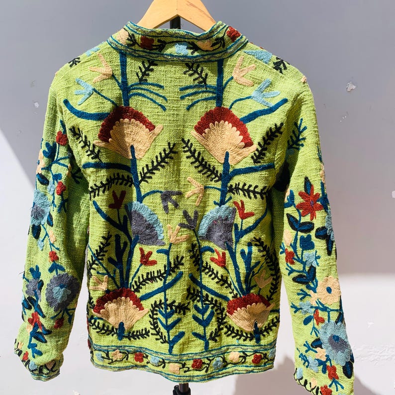 May include: A light green jacket with intricate floral embroidery in shades of red, blue, tan, and gray. The jacket features a high collar and long sleeves, with the floral design covering the front, back, and sleeves. The jacket is hanging on a wooden hanger.