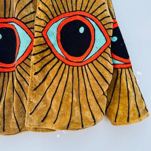May include: Mustard-colored jacket with large embroidered eye designs. The eyes feature black pupils, turquoise irises, and red outlines. Black lines radiate from the eyes, creating a sunburst effect. The jacket's fabric has a soft, velvety texture.