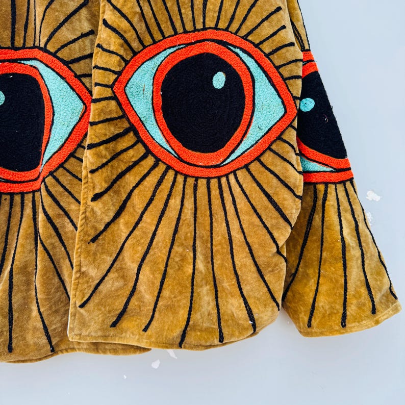 May include: Mustard-colored jacket with large embroidered eye designs. The eyes feature black pupils, turquoise irises, and red outlines. Black lines radiate from the eyes, creating a sunburst effect. The jacket's fabric has a soft, velvety texture.