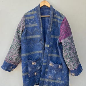 May include: A long-sleeved jacket featuring a deep blue base with intricate white and gold geometric patterns. The garment includes two front pockets and contrasting purple and green patterned fabric on the sleeves, all finished with visible running stitch detailing.