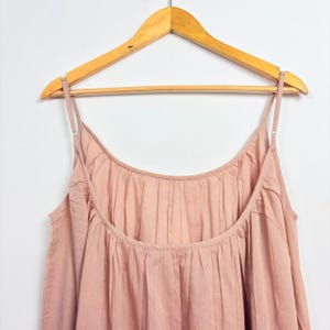 May include: A dusty rose-colored camisole dress with thin spaghetti straps, hanging on a wooden hanger. The dress features a gathered neckline and a flowing, pleated design. The fabric appears soft and lightweight, suitable for warm weather.