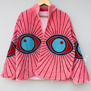 Peut inclure: Veste en velours rose ornée d'un motif d'œil. Les yeux brodés en bleu, rouge et marine sont entourés de lignes rayonnantes. Col montant et doublure rayée. Vêtement original et artistique.