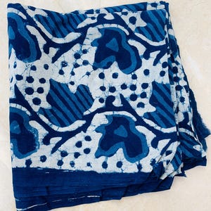 May include: Folded indigo blue and white fabric with a repeating pattern of organic shapes and dots. The fabric has a dark blue border and a fringed edge. The design is reminiscent of batik or block printing.
