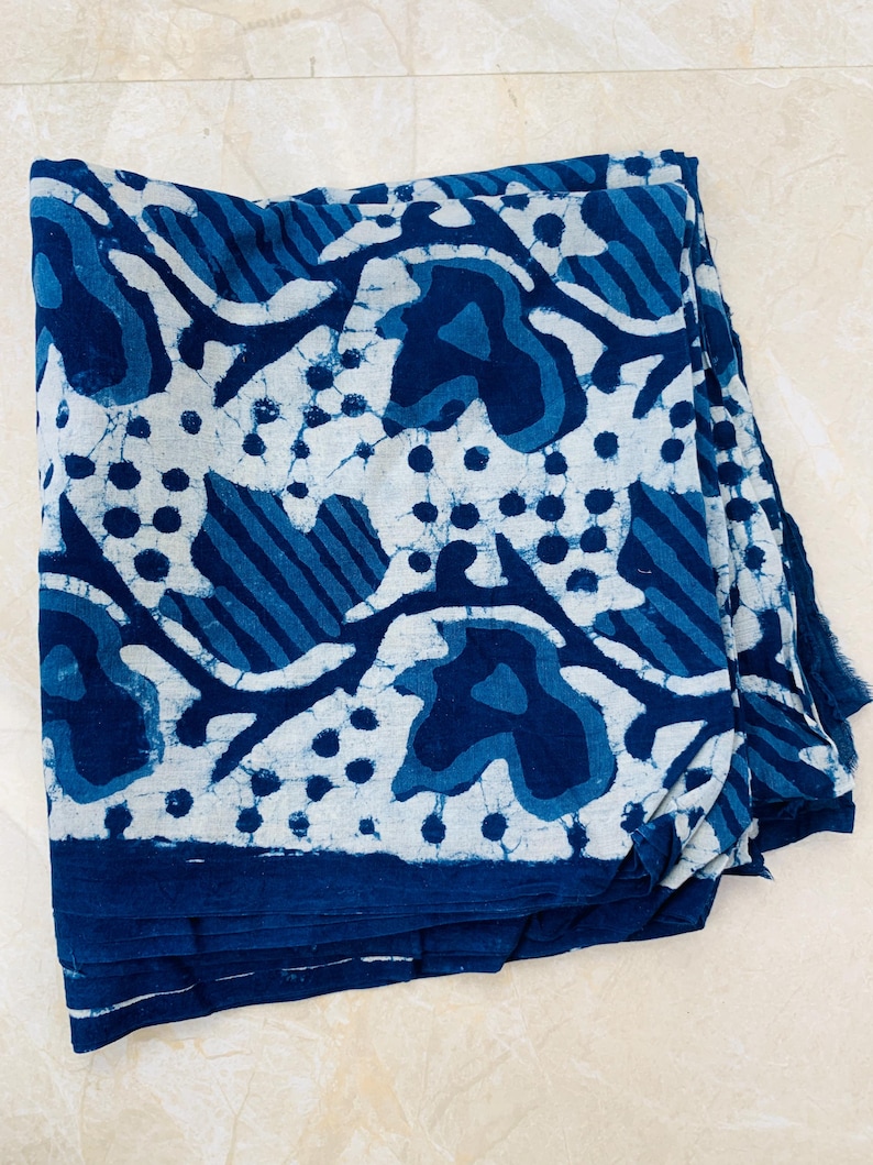 May include: Folded indigo blue and white fabric with a repeating pattern of organic shapes and dots. The fabric has a dark blue border and a fringed edge. The design is reminiscent of batik or block printing.