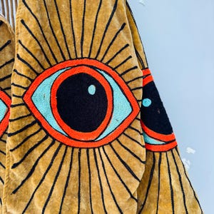May include: Close-up of a mustard-colored garment with large embroidered eye designs. The eyes feature black pupils, light blue irises, and are outlined in orange. Black lines radiate from the eyes, creating a sunburst effect.