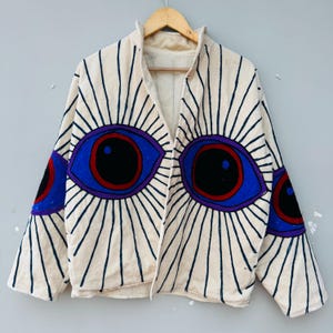 May include: Cream-colored jacket with large, embroidered eye designs in blue, purple, and black. The jacket features a collar and open front, with radiating lines emanating from the eyes. The sleeves also have eye designs.
