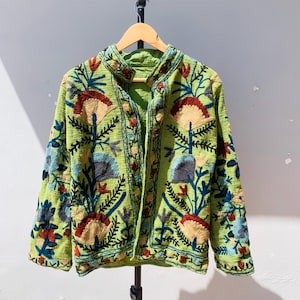 May include: A green embroidered jacket with a floral design. The jacket features a mandarin collar and long sleeves. The embroidery includes red, blue, and tan flowers and leaves. The jacket is hanging on a wooden hanger.