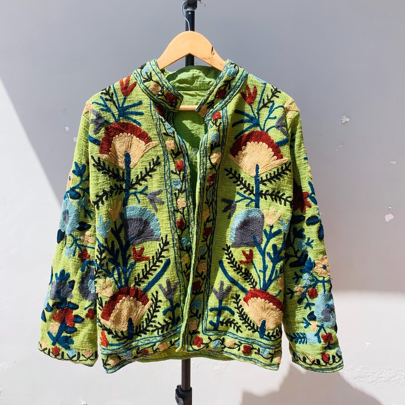 May include: A green embroidered jacket with a floral design. The jacket features a mandarin collar and long sleeves. The embroidery includes red, blue, and tan flowers and leaves. The jacket is hanging on a wooden hanger.
