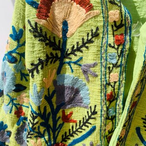 May include: Close-up of a lime green jacket with intricate floral embroidery. The embroidery features a variety of colors, including blue, red, yellow, and gray. The jacket has a textured fabric and a detailed design.