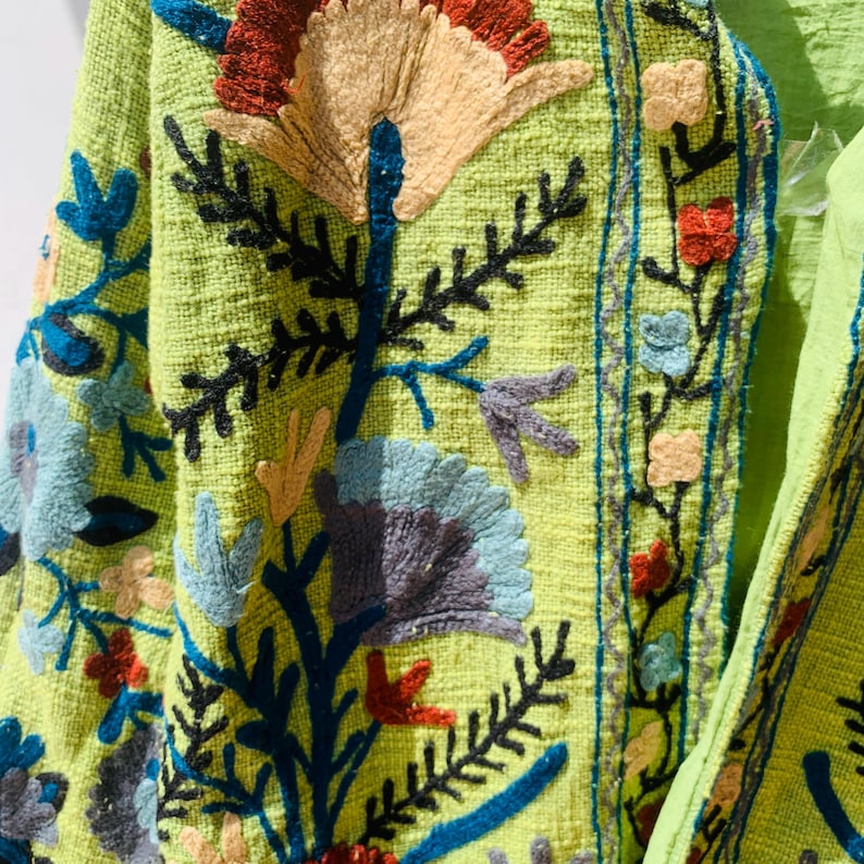May include: Close-up of a lime green jacket with intricate floral embroidery. The embroidery features a variety of colors, including blue, red, yellow, and gray. The jacket has a textured fabric and a detailed design.
