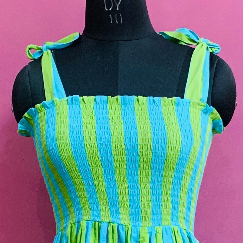 Puede incluir: Vestido sin mangas con estampado de rayas azules y verdes. El vestido presenta tirantes anudados y un corpi&ntilde;o fruncido. El vestido se exhibe en un maniqu&iacute; negro sobre un fondo rosa.