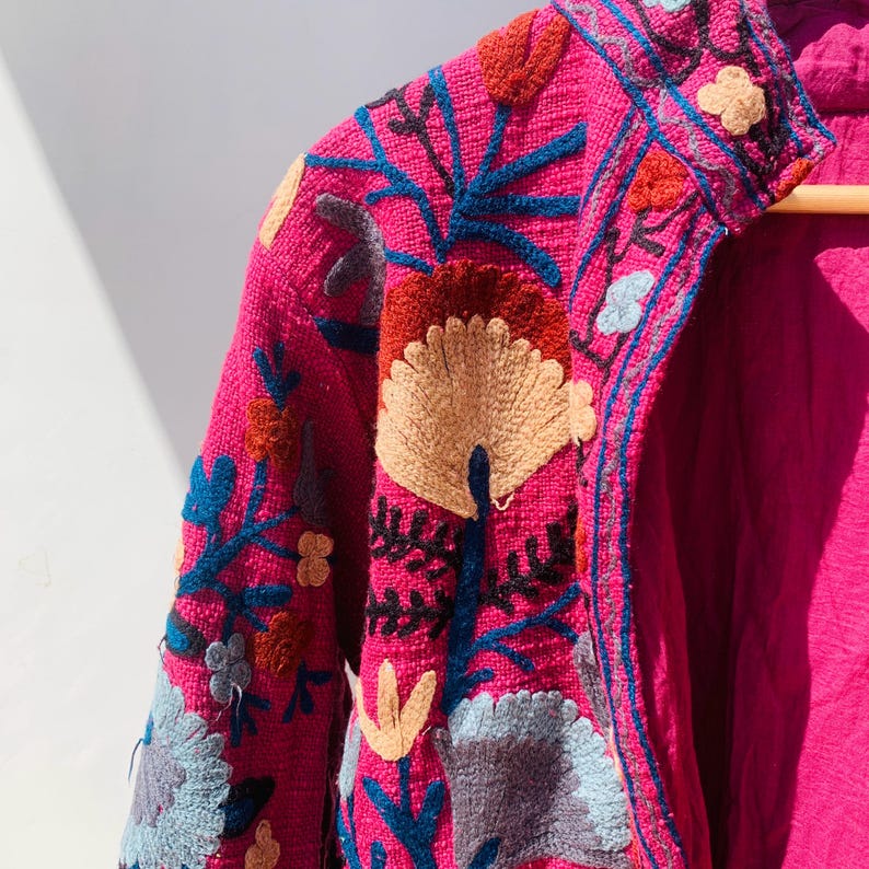Op de afbeelding: Een close-up van een magenta jasje met gedetailleerde bloemenborduursels. De borduursels bevatten blauw, oranje, beige en zwart, waardoor een textuurontwerp ontstaat. De jas is gemaakt van een geweven stof. De maten zijn in centimeters.