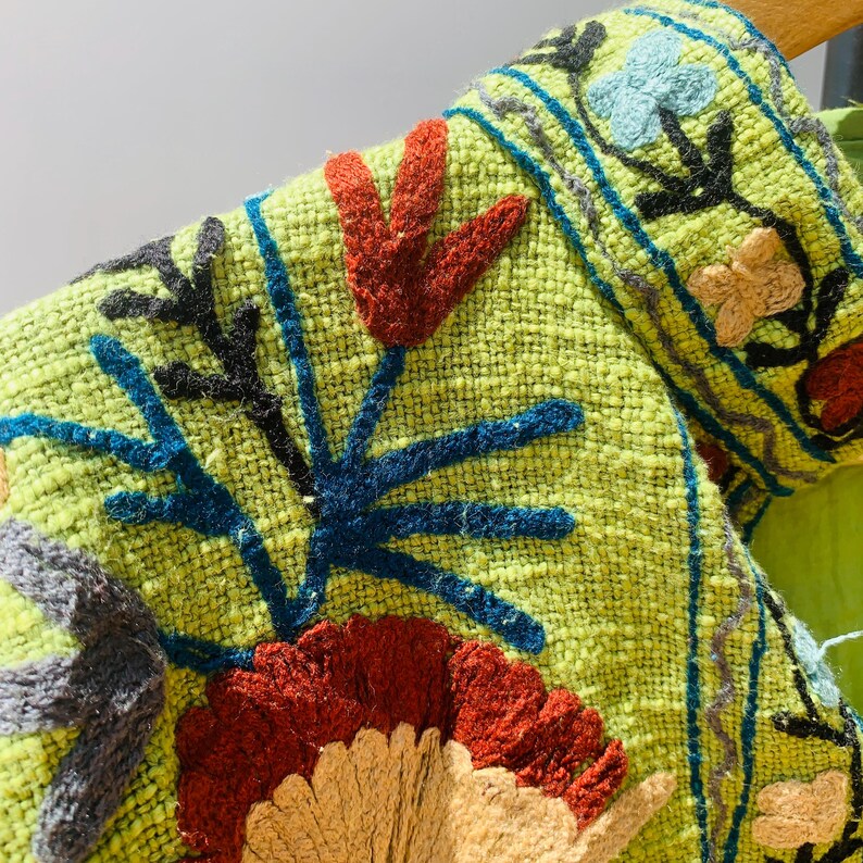 May include: Close-up of a lime green textile with colorful embroidered floral designs. The embroidery features red, blue, black, and tan flowers and leaf patterns. The fabric has a woven texture.